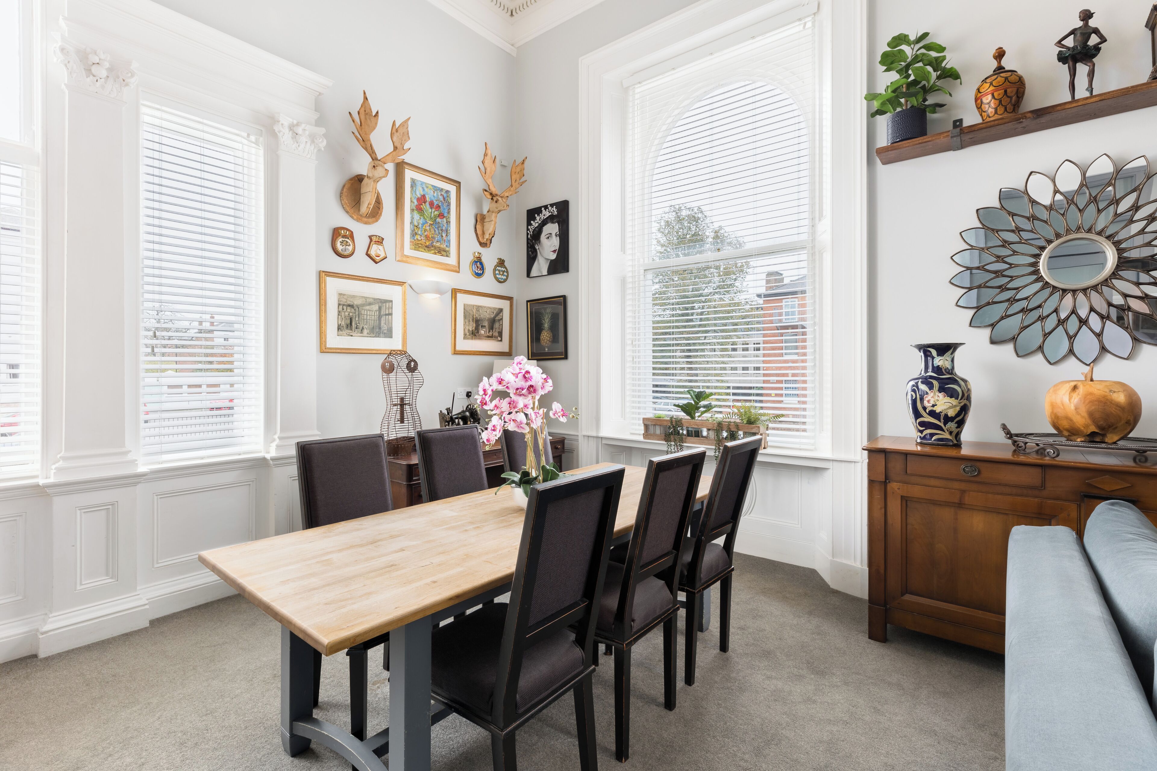 Un comedor con mesa y sillas de madera, rodeado de amplios ventanales con persianas blancas.