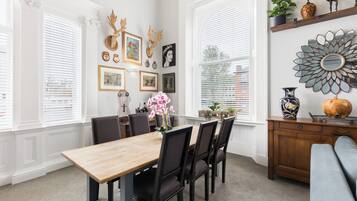 Un comedor con mesa y sillas de madera, rodeado de amplios ventanales con persianas blancas.
