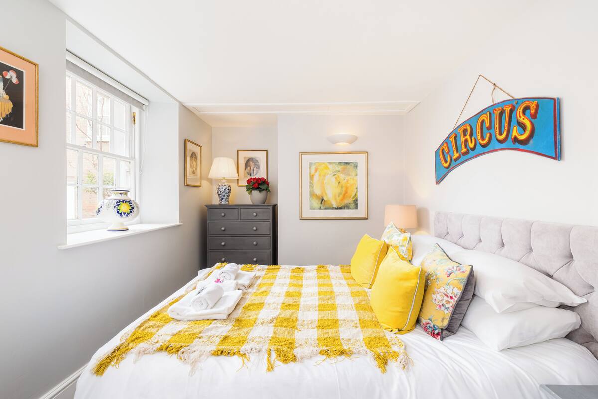 A bedroom with a checkered bedspread, a 'Circus' sign, a window with a view, and a dresser with a vase of flowers.