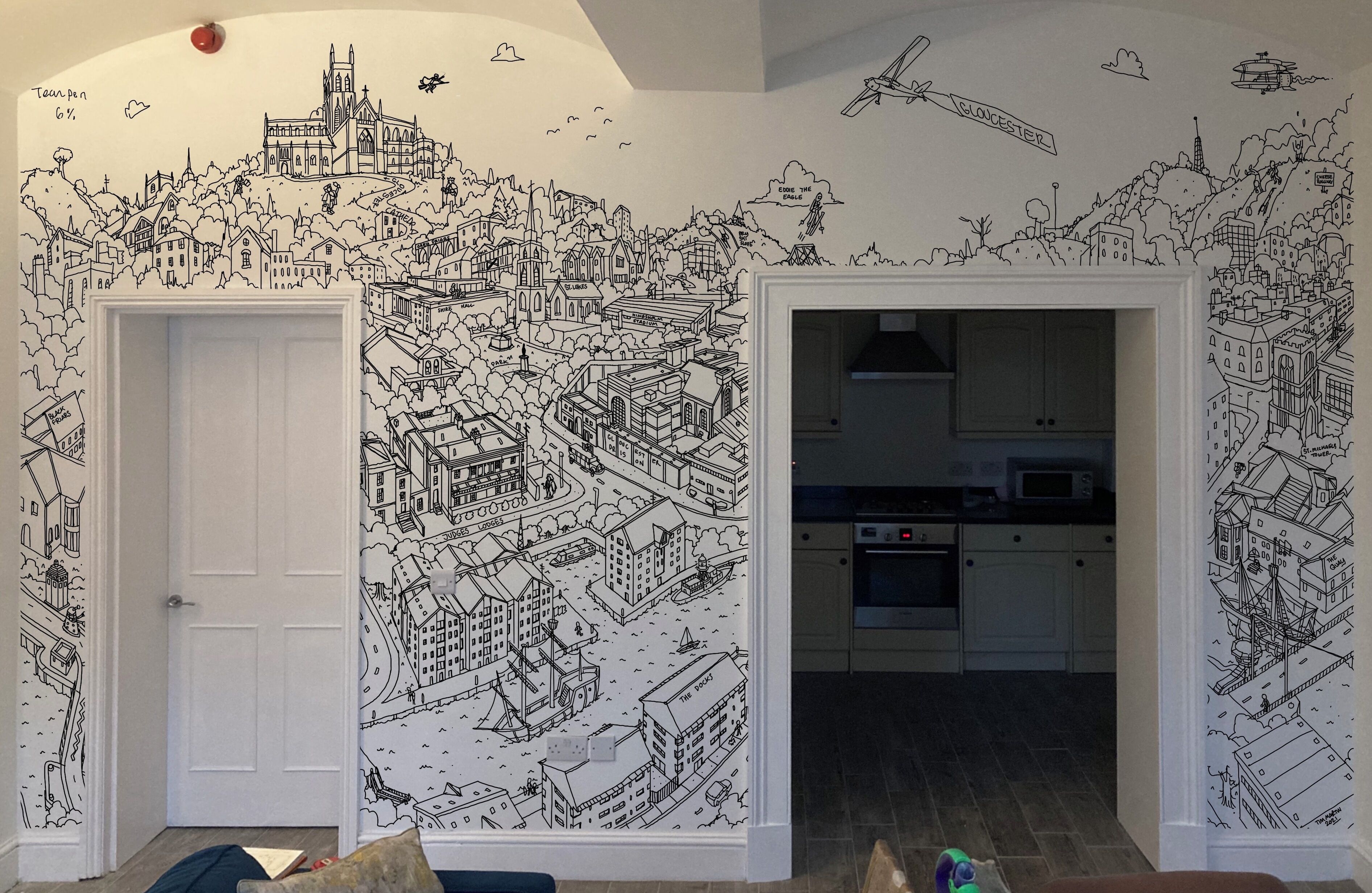 Habitació amb mural de paisatge urbà a les parets, una porta blanca i una zona de cuina visible a través d'una obertura.