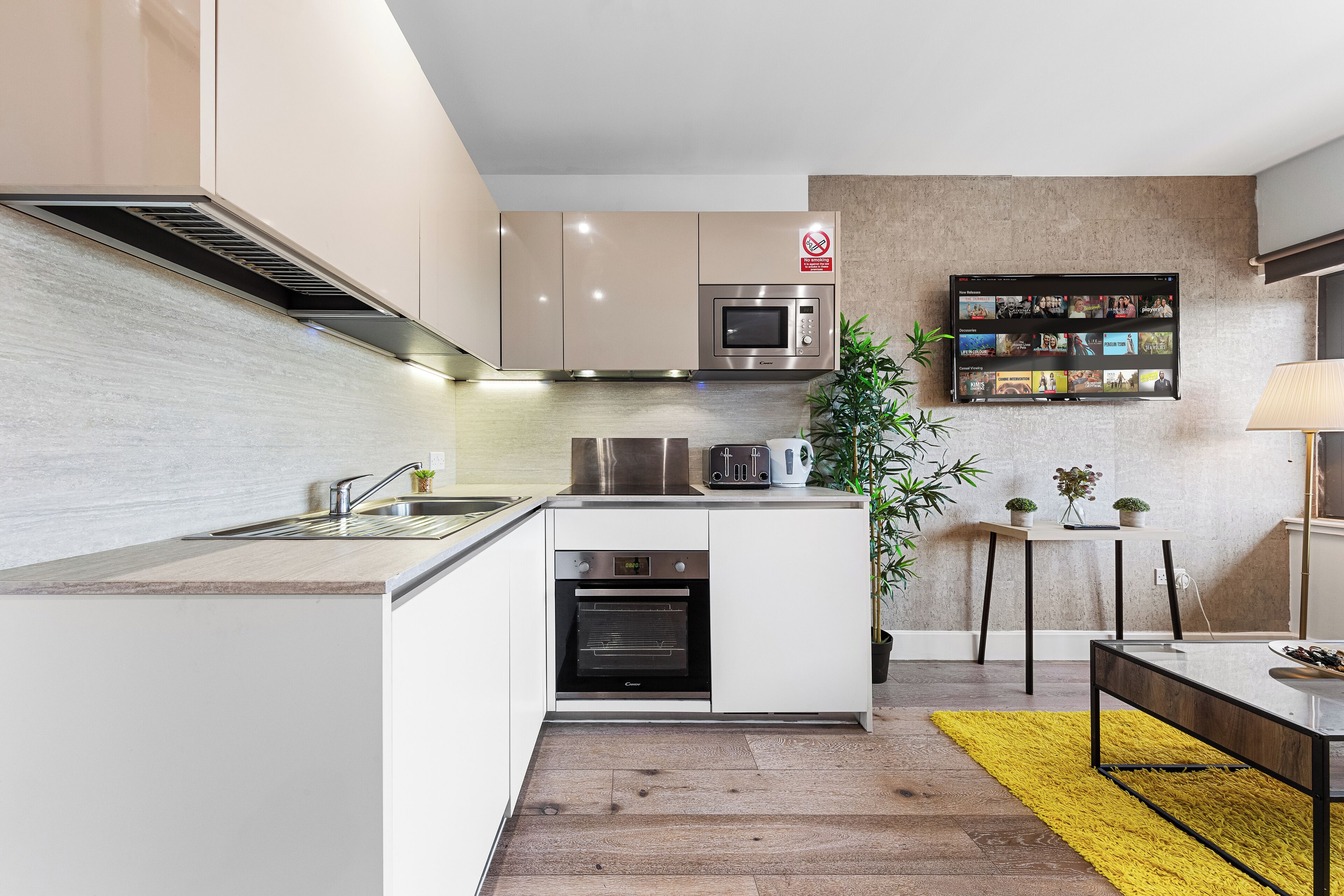 Una cocina moderna con electrodomésticos integrados, un comedor con un tapete amarillo y un televisor de pared mostrando varios canales.