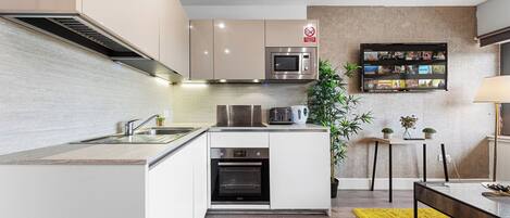 Una cocina moderna con electrodomésticos integrados, un comedor con un tapete amarillo y un televisor de pared mostrando varios canales.