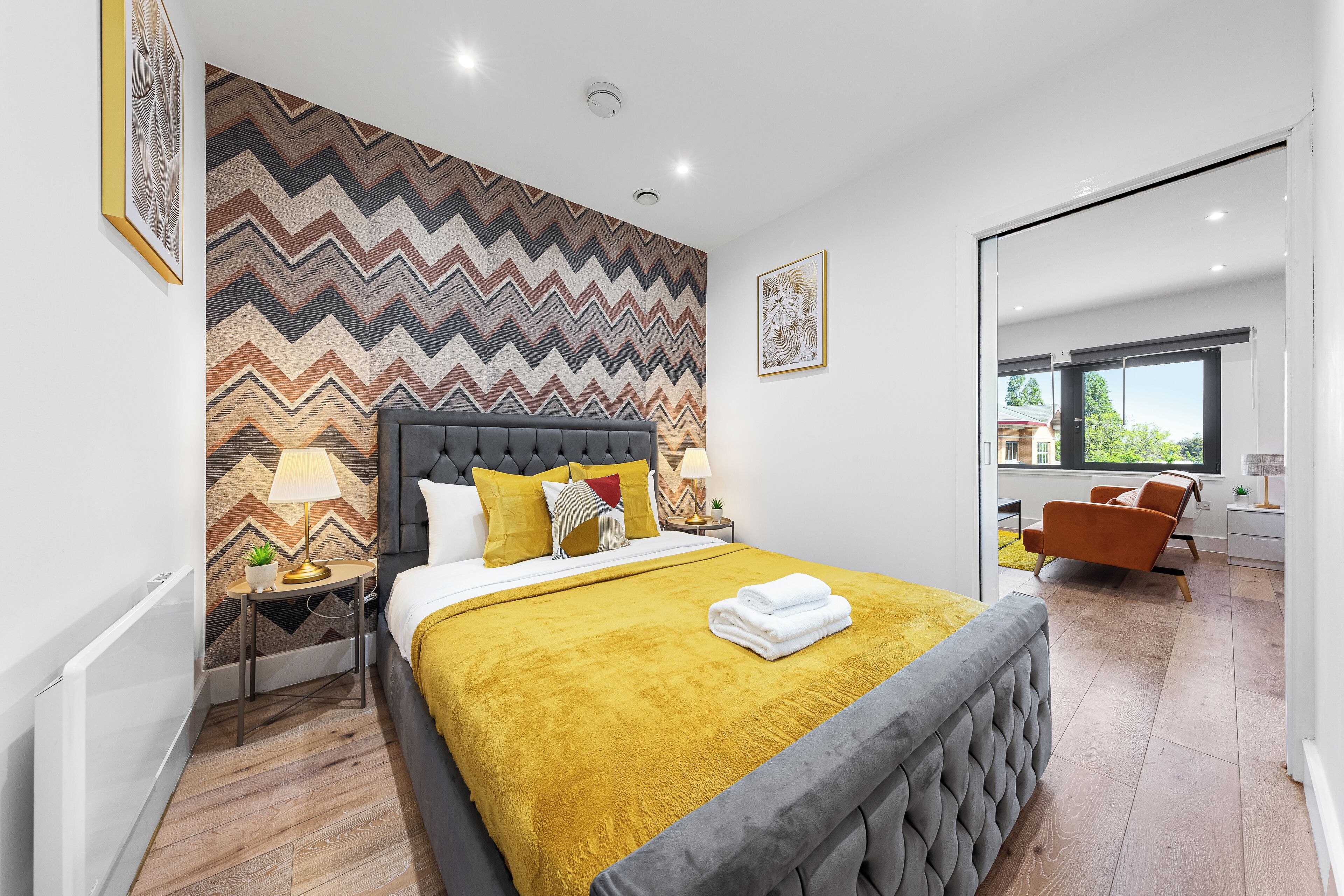 A modern hotel room with a large bed, a chevron-patterned wall, a bedside table with a lamp, and a view of the outdoors.