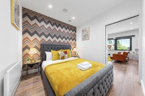 Una habitación de hotel moderna con una cama grande, una pared con estampado en zigzag, una mesita de noche con lámpara y vistas al exterior.