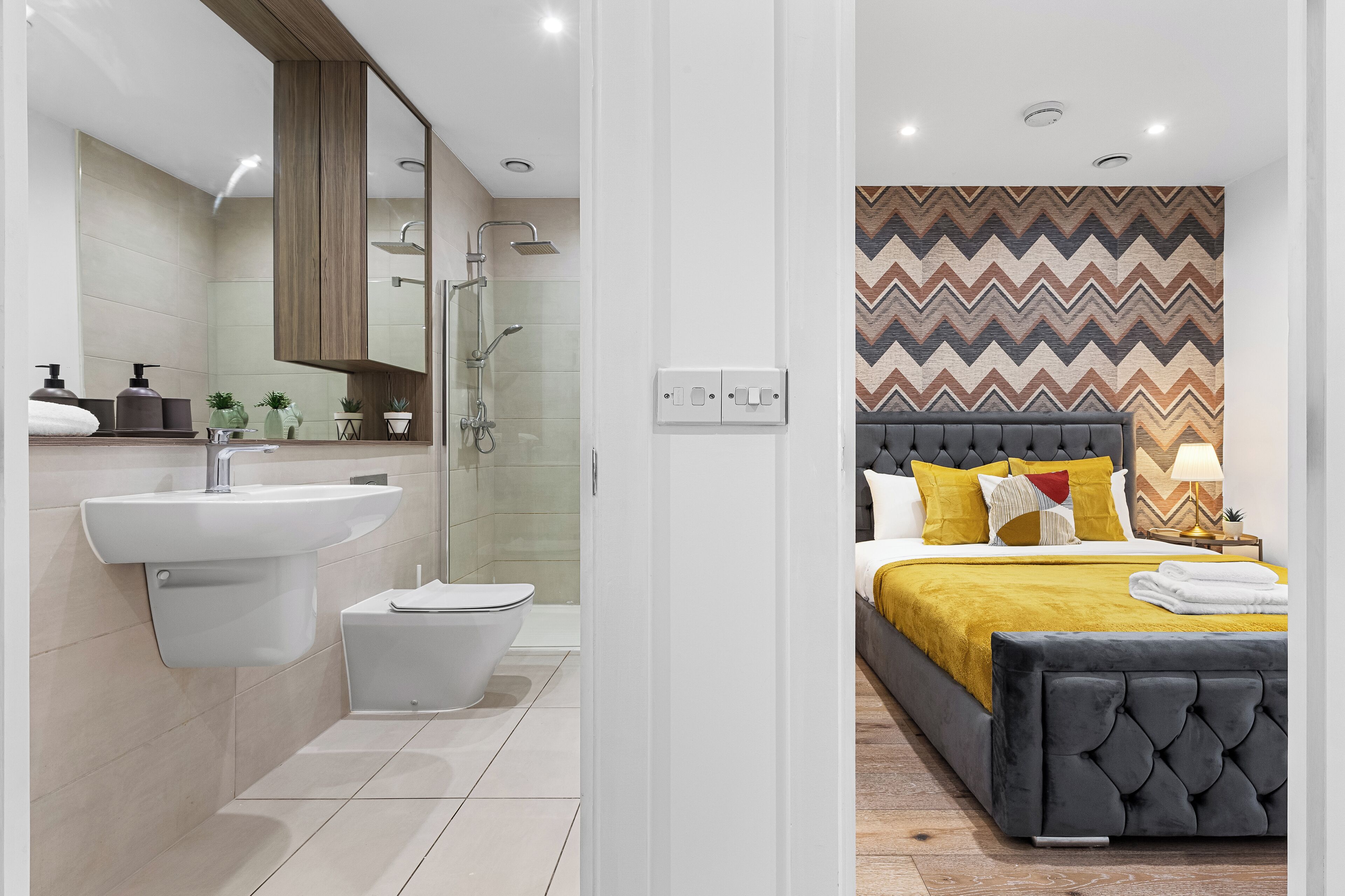 A modern bathroom with a sink, toilet, and shower. The room features a large mirror, a patterned wall, and a bed with a black headboard and yellow bedding.