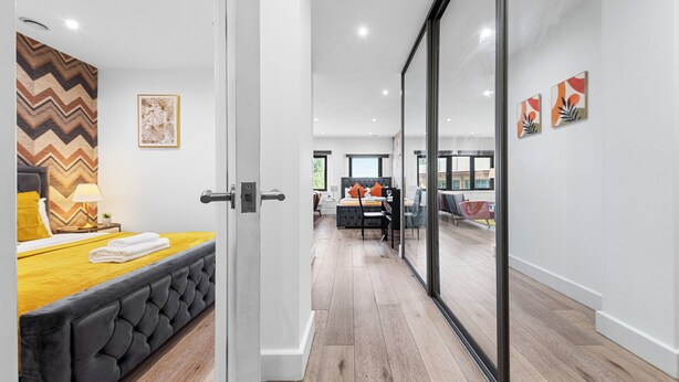 A modern hotel room with a large bed, a wooden headboard, a mirror, and a view of the adjacent living area through a glass door. - Hayes Haven - Charming 1 Bedroom Apartment (Hayes)