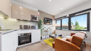 A modern kitchen with white cabinets, a built-in oven, and a microwave. There's a dining area with a yellow rug and orange chairs. - Hayes Haven - Charming 1 Bedroom Apartment (Hayes)