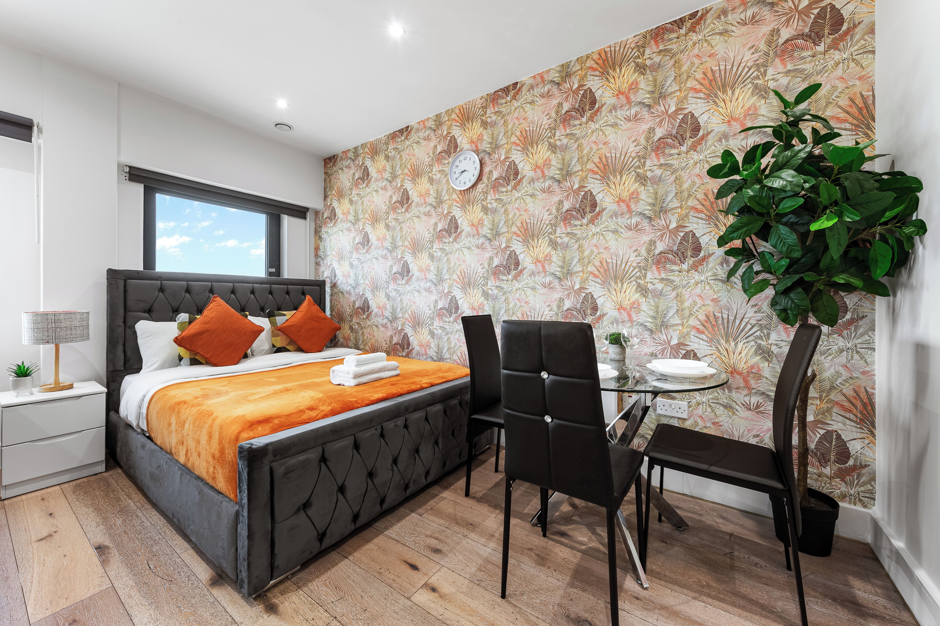 A modern bedroom with a tufted headboard bed, a bedside table with a lamp, a large window, a dining area with a table and chairs, and a wall with a tropical leaf pattern.