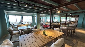 An indoor dining area with wooden tables and chairs, large windows offering views of the outdoors, and a high ceiling with exposed beams. - Mt. Plaisir Estate Hotel (Grande Riviere)