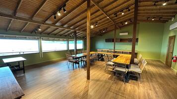 A spacious indoor area with wooden tables and chairs, large windows, and a high ceiling with exposed beams.