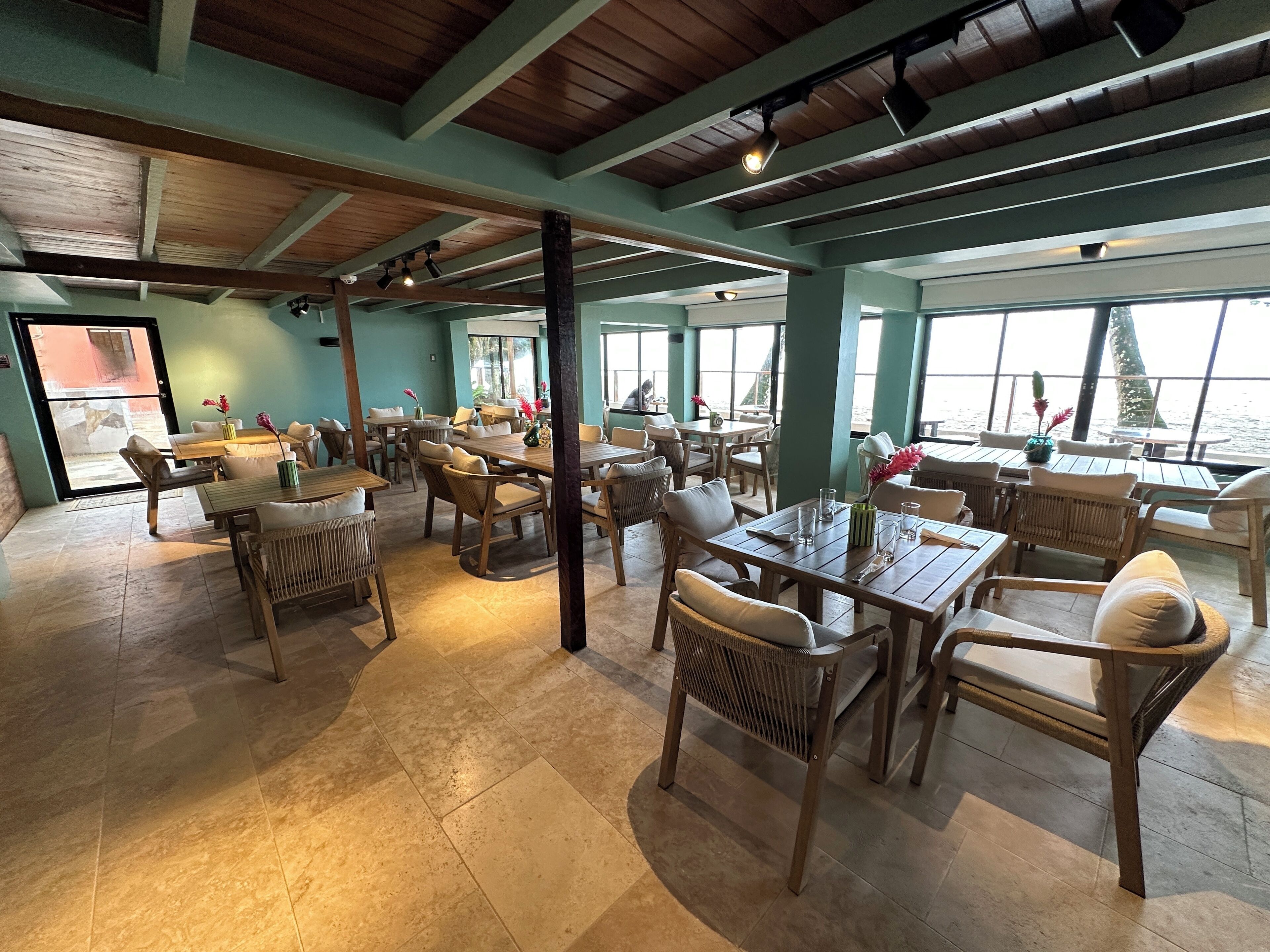 A spacious dining area with wooden tables and chairs, large windows offering views of the sea, and a high ceiling with exposed beams.