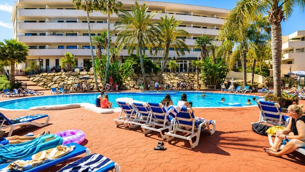 A pool area with lounge chairs and palm trees, surrounded by a building with multiple floors. - Wake up to pool views · Festival Village (Salou)