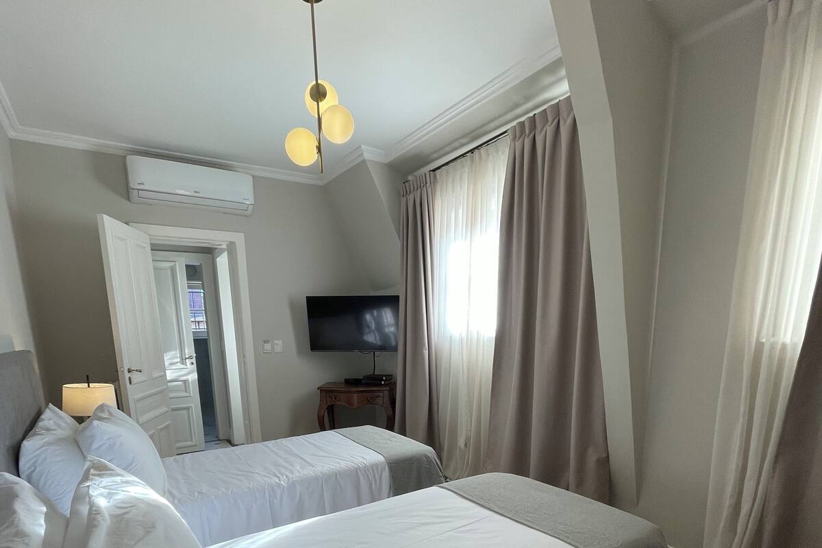 A hotel room with two beds, a television, an air conditioning unit, and a door leading to another room.