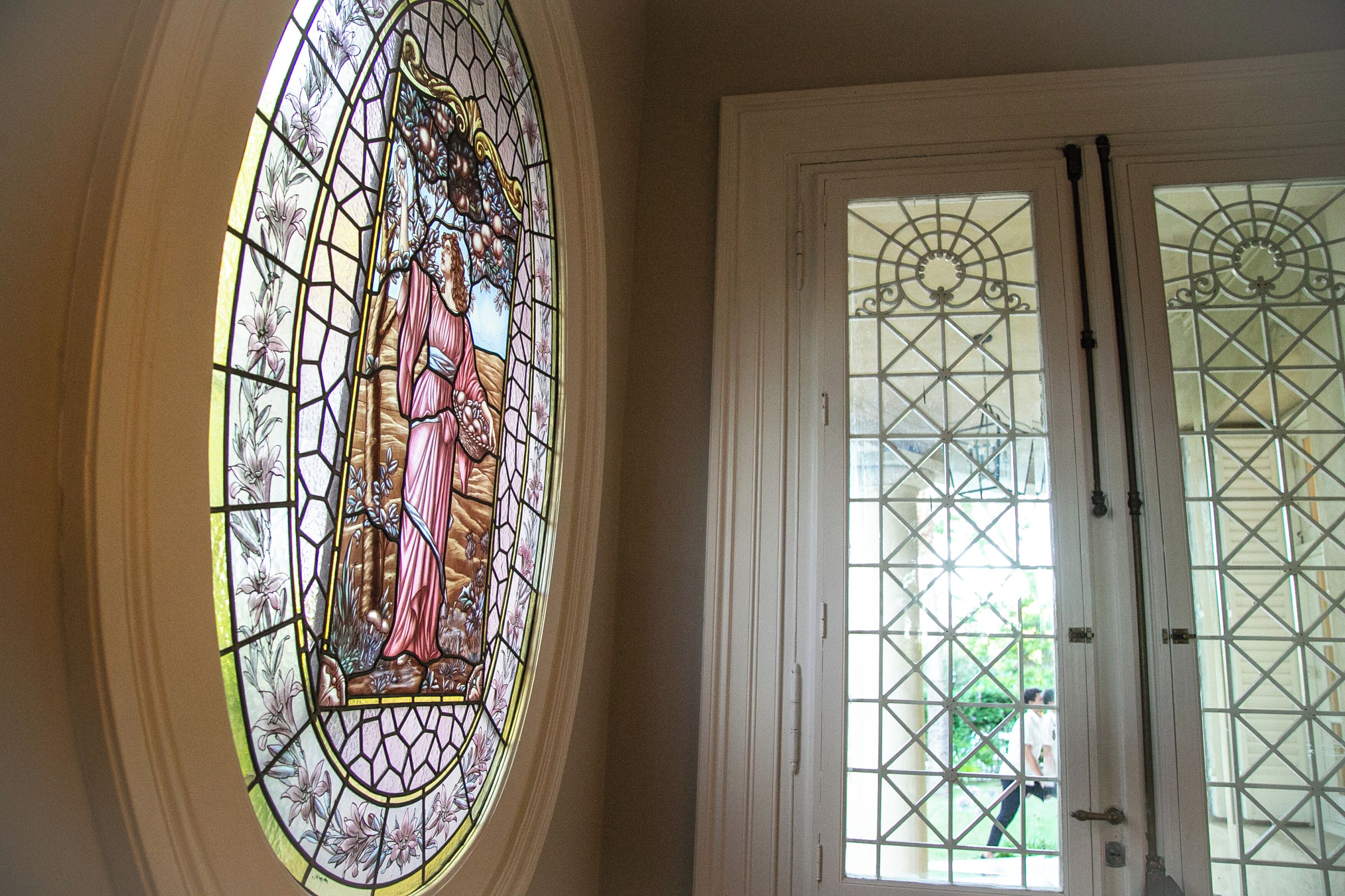 A stained glass window with a religious figure, a white door with a metal grid, and a view of greenery outside.