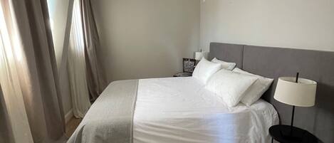 A neatly made bed with white linens, a grey headboard, and a bedside table with a lamp.