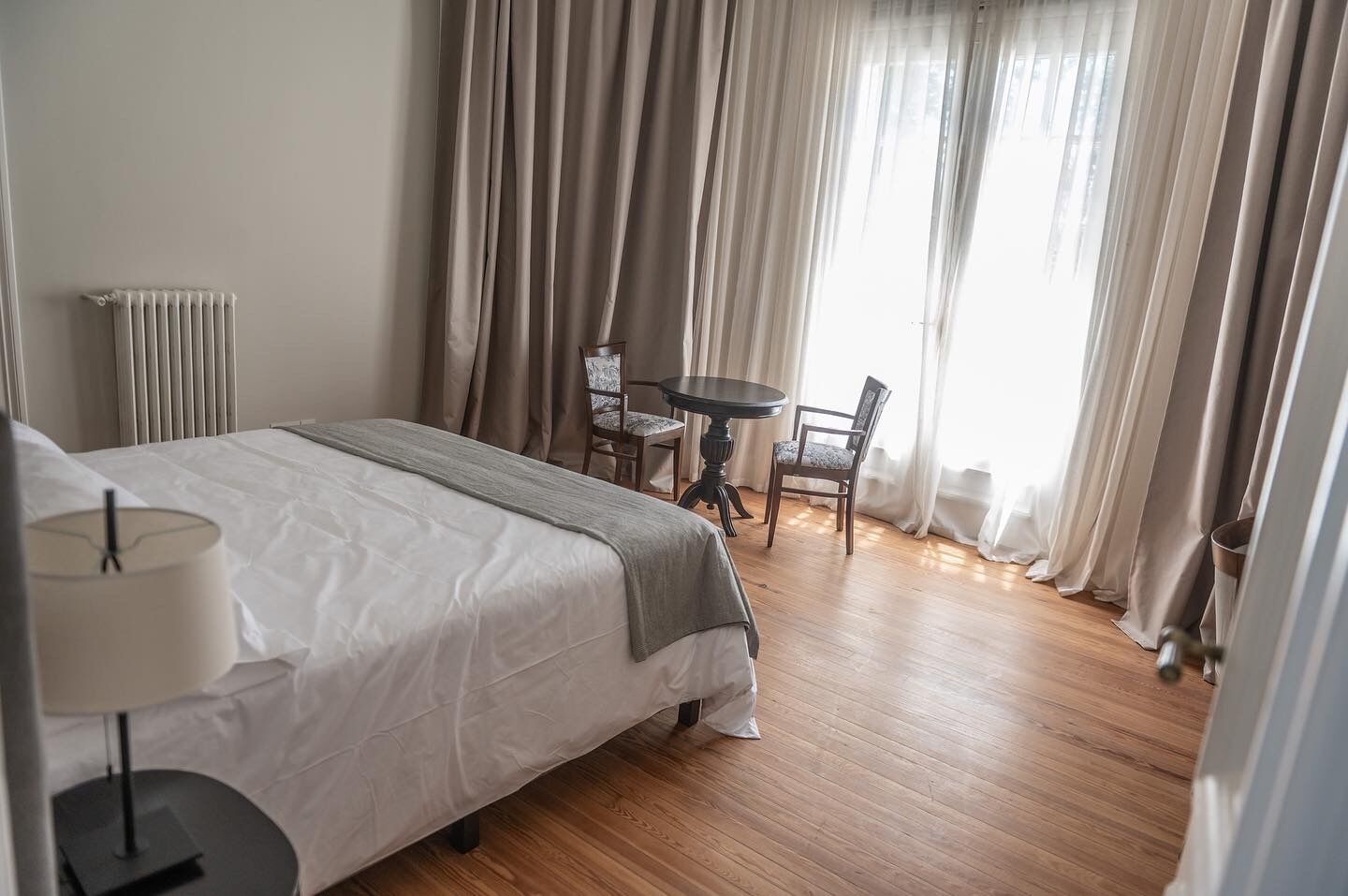 Quarto de hotel com cama, mesa, duas cadeiras, luminária e janela com cortinas.