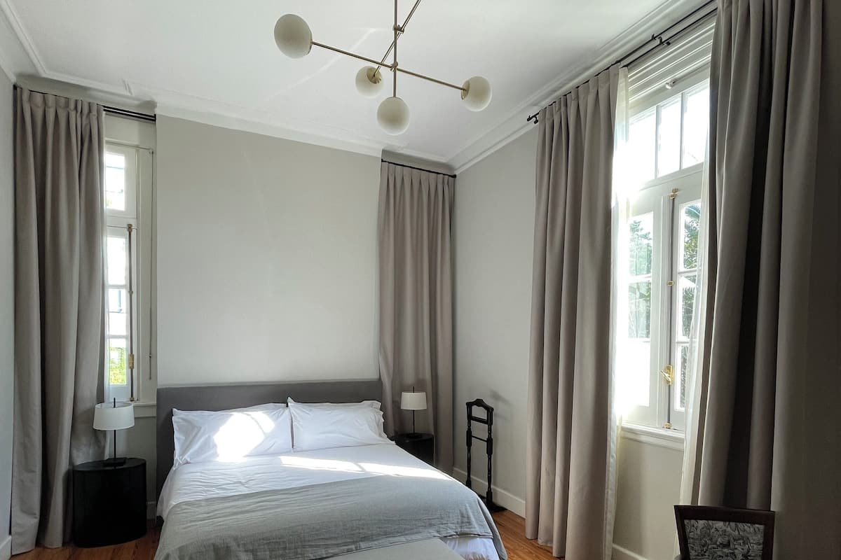 A bedroom with a large bed, a bedside table, a chair, a lamp, and a window with curtains.