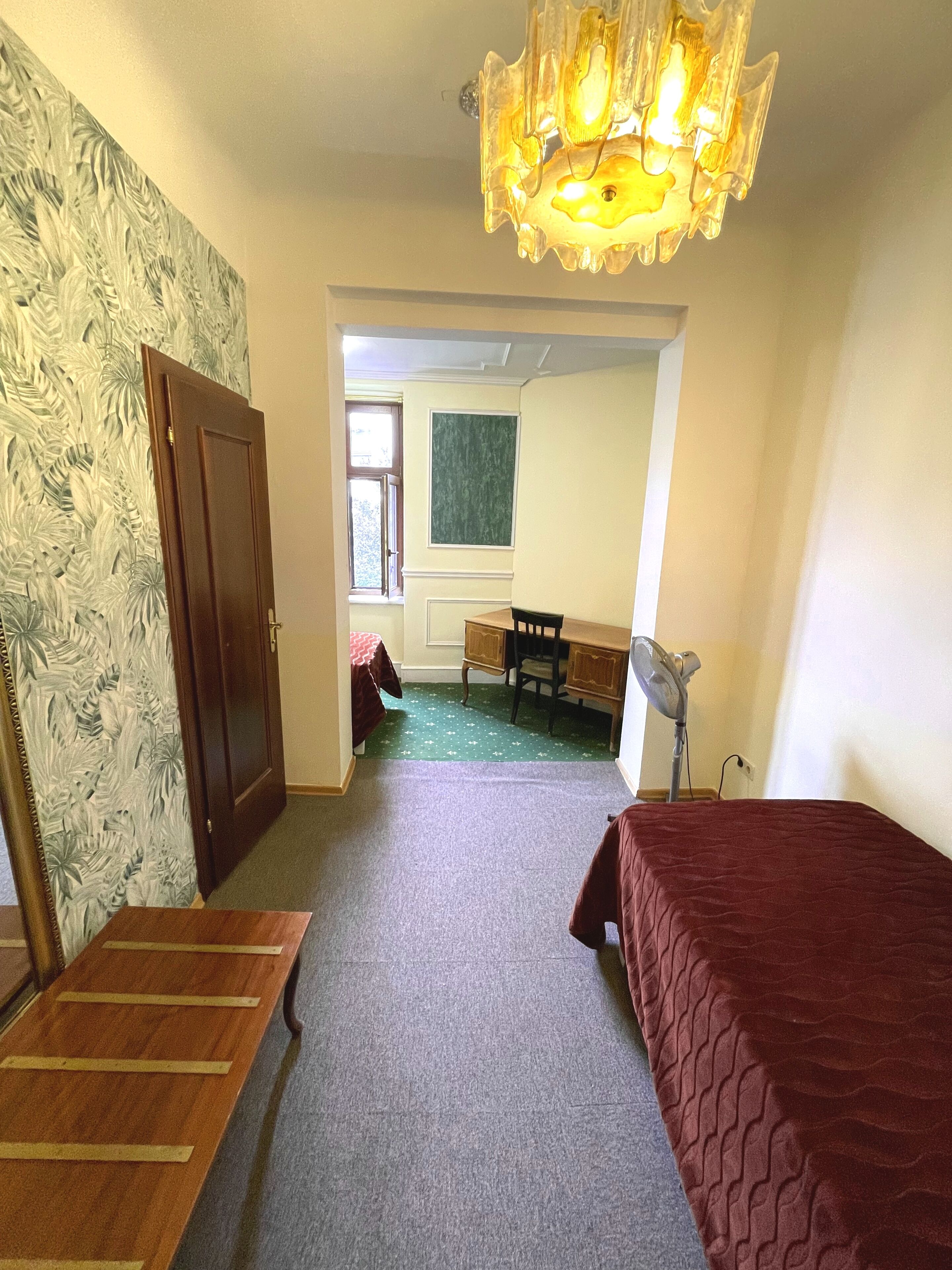 Una habitación con un banco de madera, una cama, un escritorio y una silla. Hay una puerta a la izquierda y una ventana a la derecha.