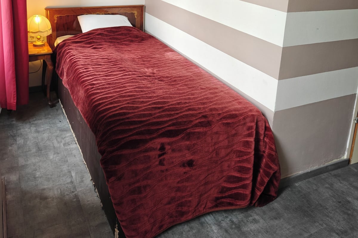 Ein kleines, ordentlich bezogenes Bett mit roter Decke in einem Zimmer mit Streifenmuster an den Wänden und einem Holz-Kopfteil.