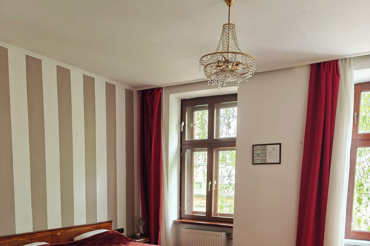 Ein Schlafzimmer mit einem hölzernen Bett, einem roten Teppich, einer Kristallleuchtre, einem Fenster mit roten Vorhängen und einem Holztisch mit einem Stuhl.