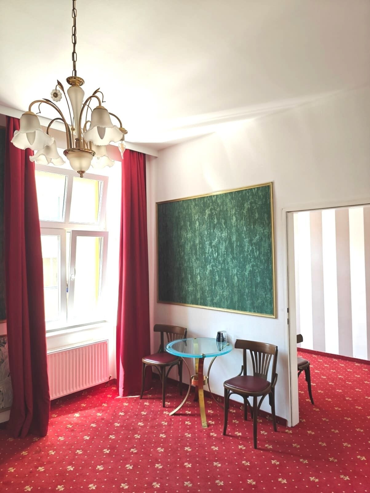 Une pièce avec un grand tableau noir vert, deux chaises en bois, une petite table ronde, un lustre et une moquette rouge.