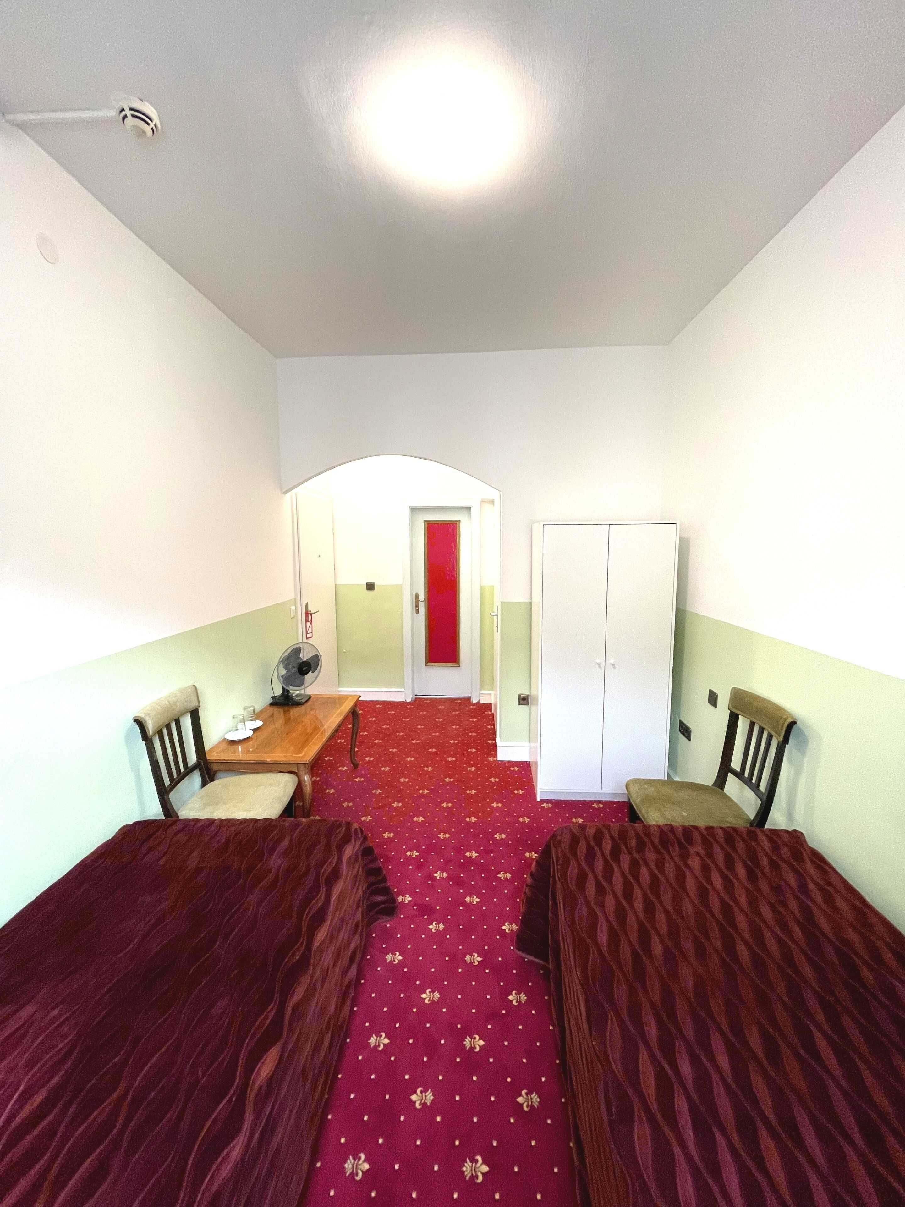 Quarto com duas camas de solteiro, uma mesinha com cadeiras, tapete vermelho e uma porta branca ao fundo.