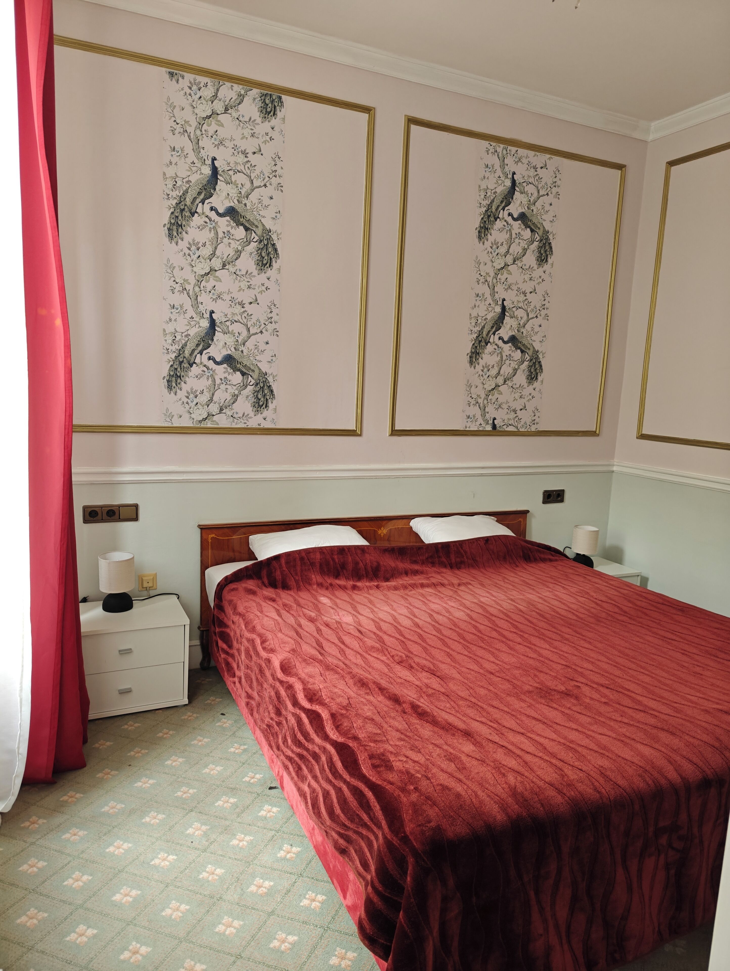 A bedroom with a large bed, a nightstand with a lamp, a red curtain, and two wall-mounted paintings of peacocks.