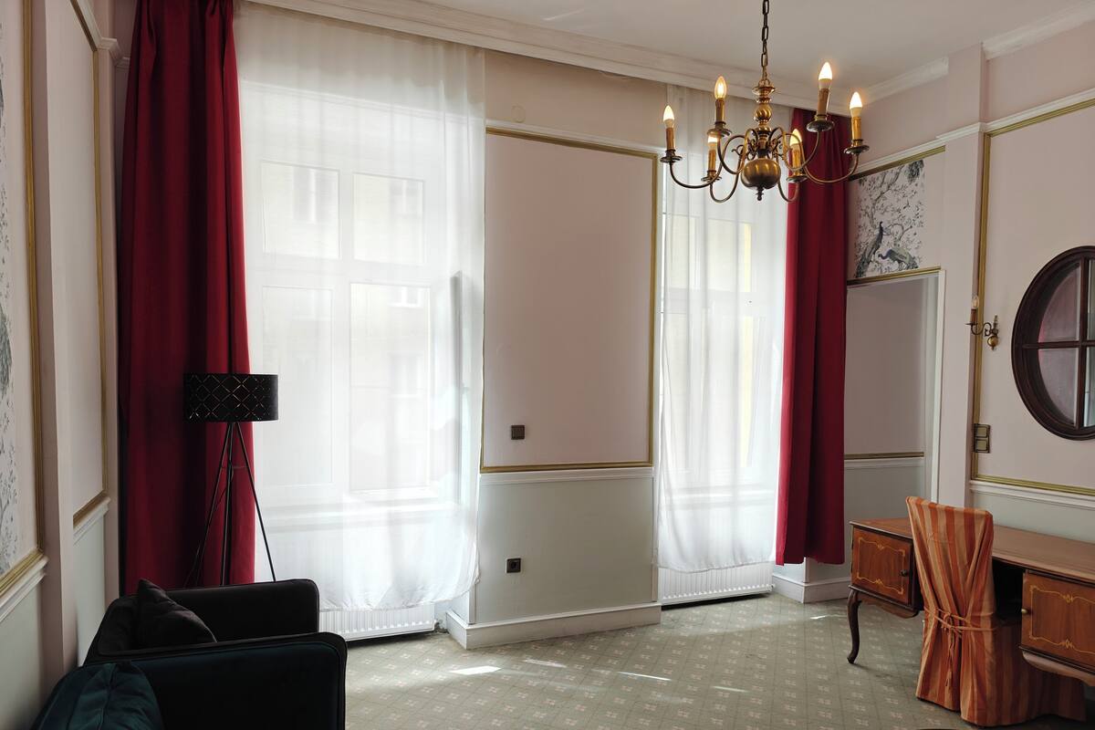 Ein Zimmer mit großem Fenster, einer Kristallleuchtre, einem Spiegel und einer Holzkommode.