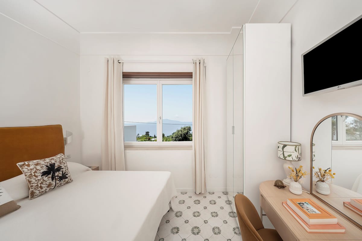 Habitación de hotel con cama, escritorio, silla, televisor, ventana con cortinas y vistas al exterior.