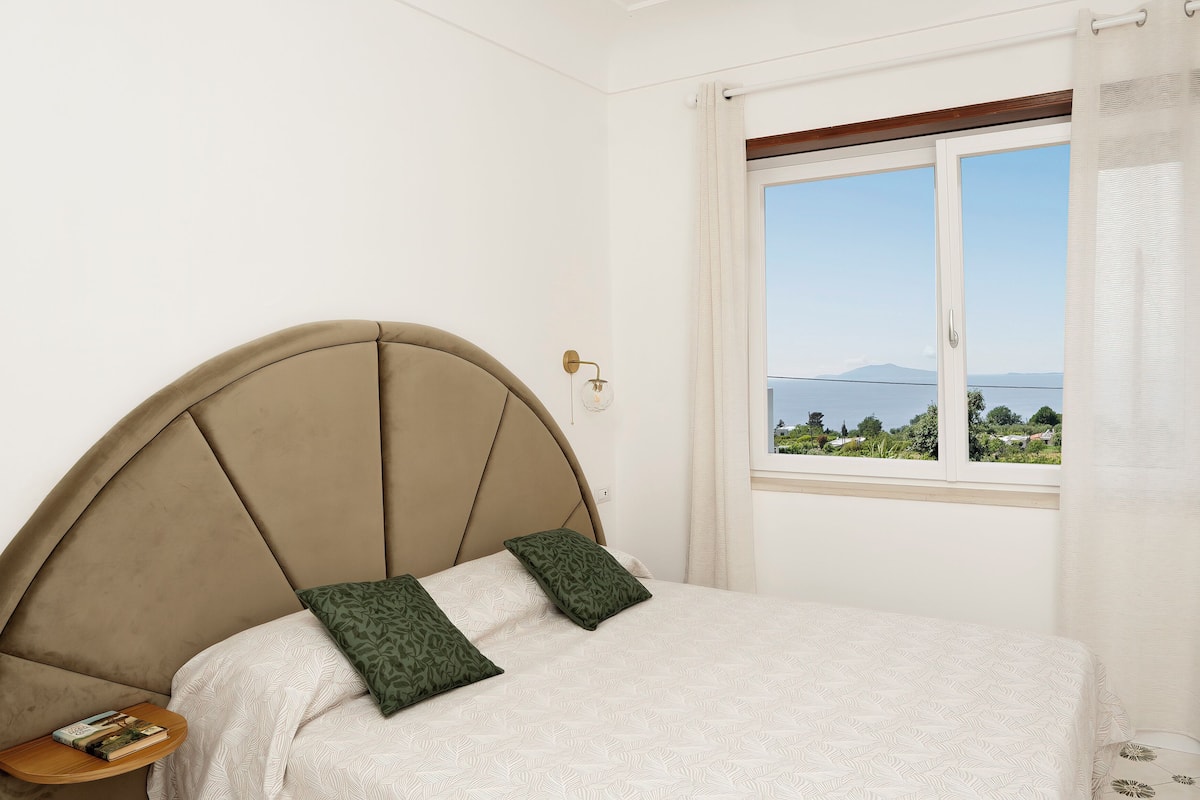 Un dormitorio con cabecera curvada, una mesita de noche con un libro, dos almohadas estampadas y vistas a una masa de agua a través de una ventana.