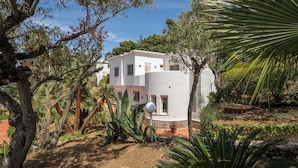 Un bâtiment moderne aux murs blancs et à la façade incurvée, entouré d’une végétation luxuriante et d’arbres.