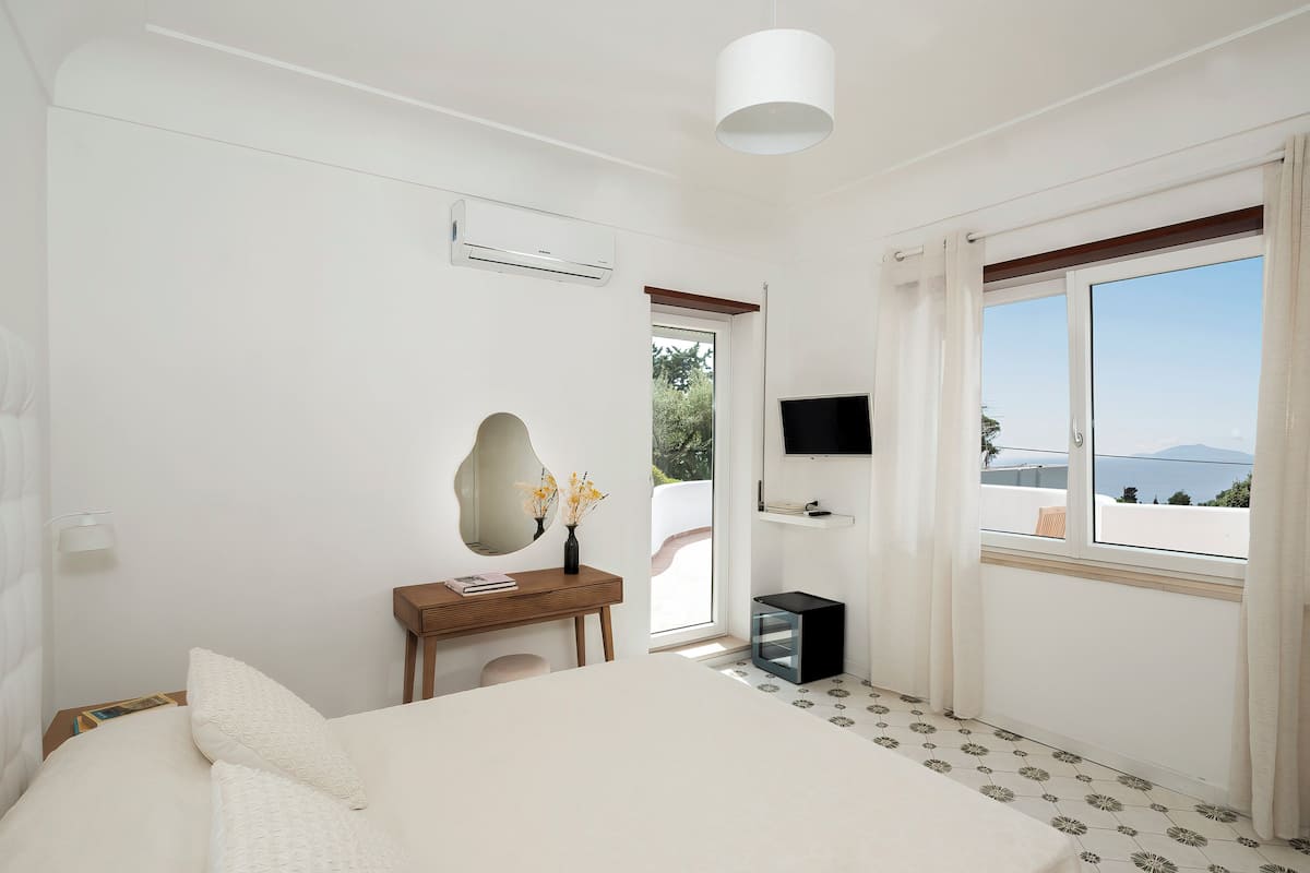 Un dormitorio con cama, mesita de noche con espejo, televisor montado en la pared y ventana con cortinas.
