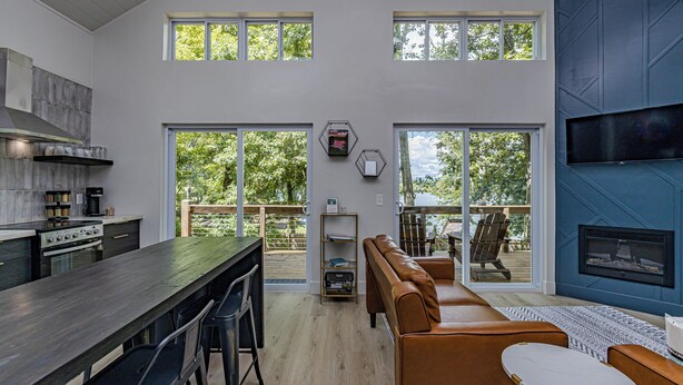 Uma cozinha espaçosa com uma ilha central, uma sala de jantar com uma mesa comprida e cadeiras, um espaço de estar com sofá e cadeiras, e uma grande porta de vidro deslizante que dá para um deck externo.