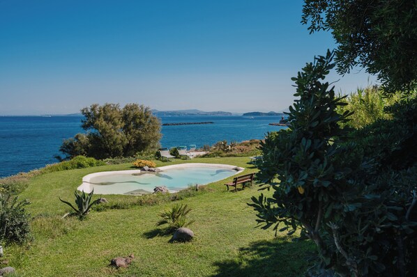 Piscina con sillones de descanso y un banco, rodeada de vegetación y con vistas al mar.