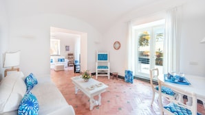 A bright living room with a white sofa, a coffee table, and a dining area with a white table and chairs. There is a large window with a view of the outdoors. - Villa Serao & Cottage Camilla by MyVillaCollec... (Ischia Porto)