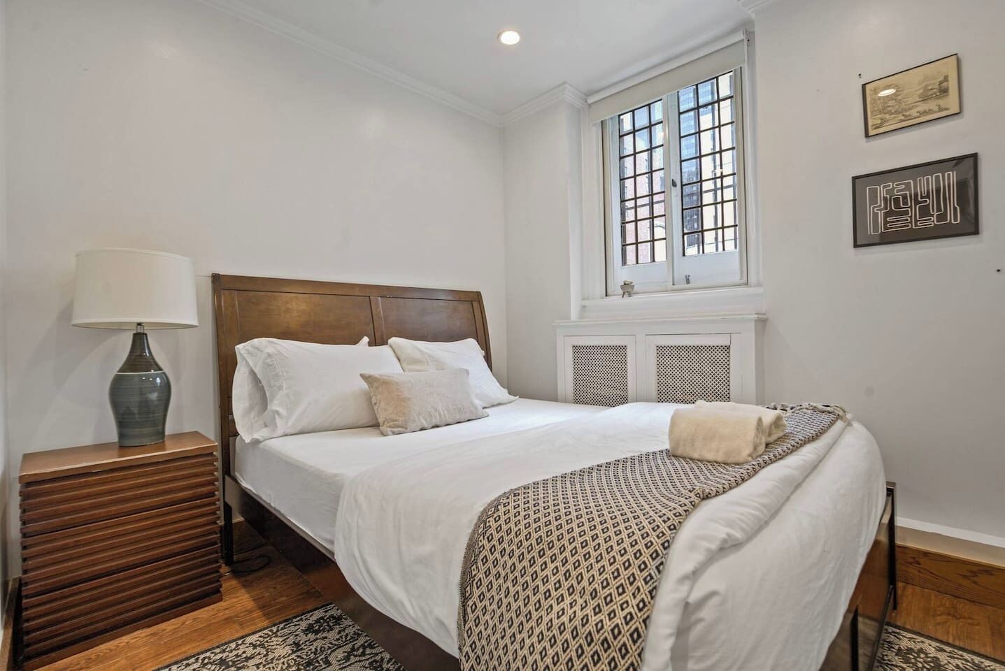 A neatly arranged bedroom with a wooden headboard, a bedside table with a lamp, a patterned throw blanket, and a window with a view.