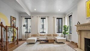 Un salon spacieux avec un grand canapé d’angle, une table basse et une cheminée. De grandes fenêtres sont agrémentées de rideaux, un escalier avec des rampes en bois est présent, et une grande fenêtre s’étend du sol au plafond sur le côté droit.