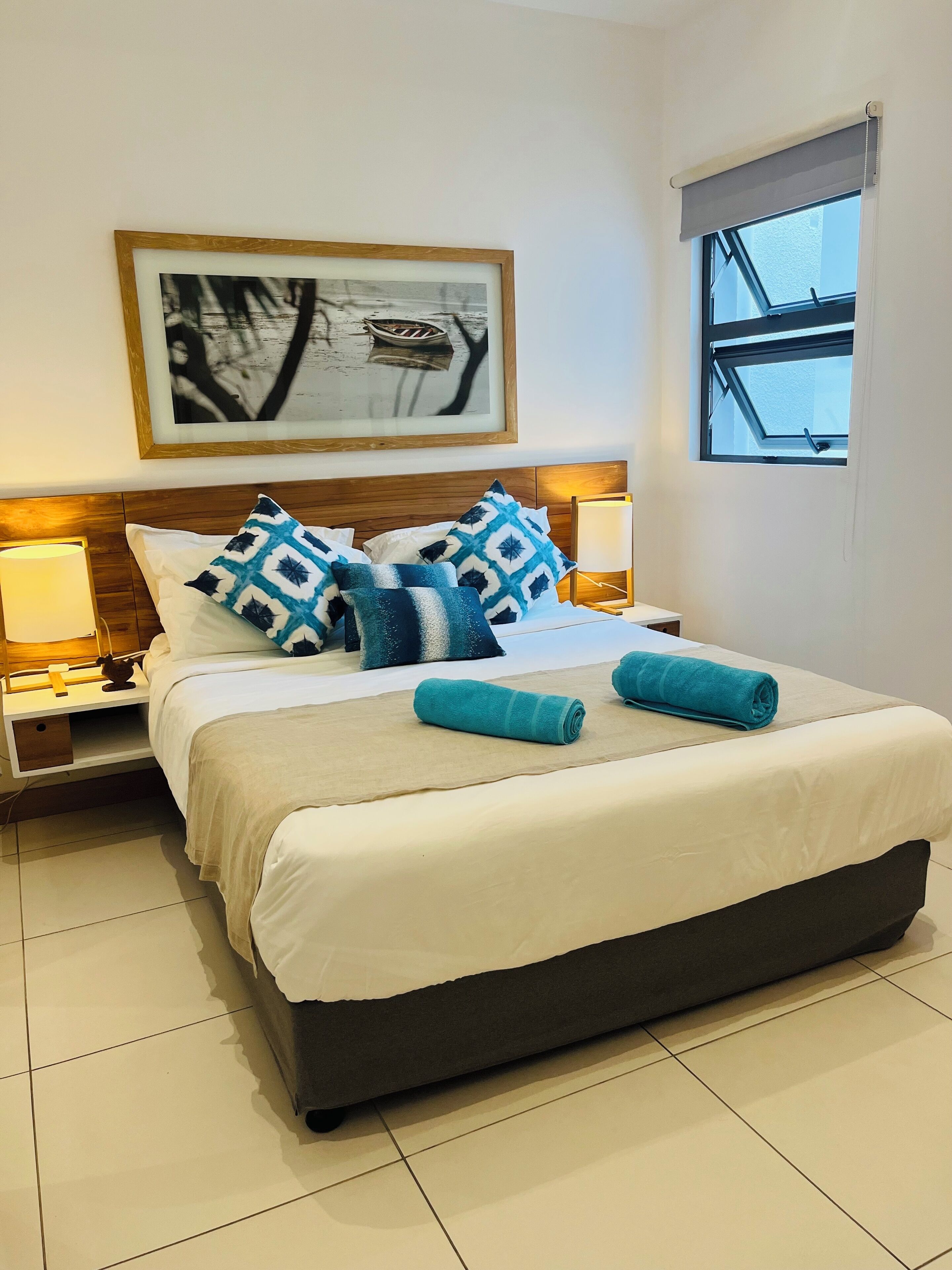 A neatly made bed with patterned pillows, a framed artwork above it, and a window with a view.