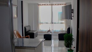 A modern living room with a kitchen area, a large window, and a flat-screen TV mounted on the wall.