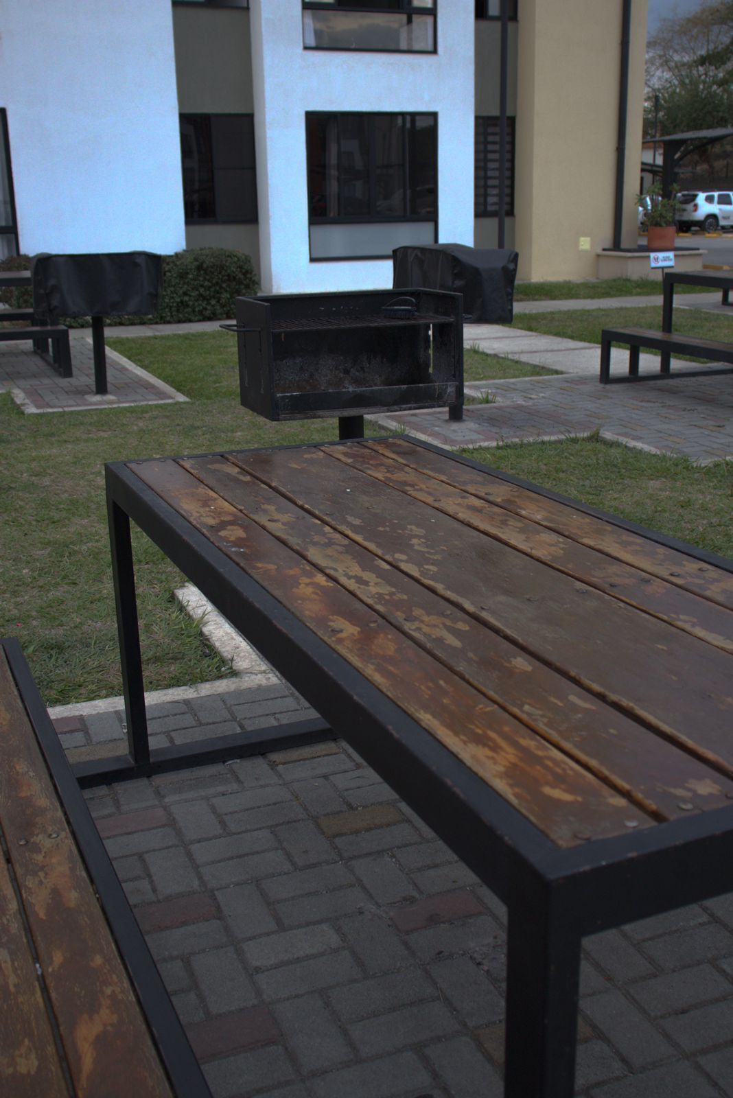 An outdoor seating area with a wooden table and benches, surrounded by a paved walkway and a building with large windows.