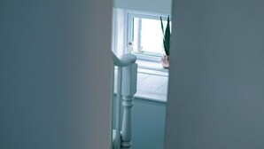 Ein schmaler Flur mit einer weißen Tür, die in einen Raum mit Fenster und Pflanze führt.