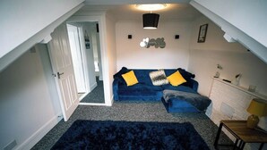 Ein Zimmer mit einem blauen Sofa, einem dunklen Teppich, einem Kamin aus weißen Ziegelsteinen und einer Deckenleuchte.