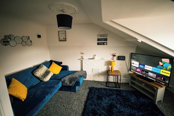 A living room with a blue sofa, a television displaying streaming services, and a white brick wall.