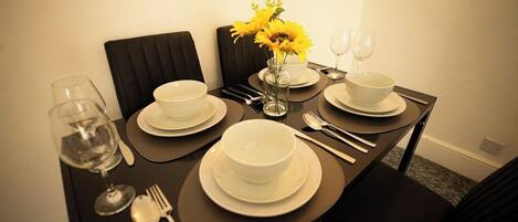 Uma mesa de jantar posta com pratos, tigelas, talheres e copos de vinho brancos, com um vaso de flores amarelas ao centro.