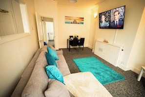 A living room with a sofa, a dining table, and a TV mounted on the wall.