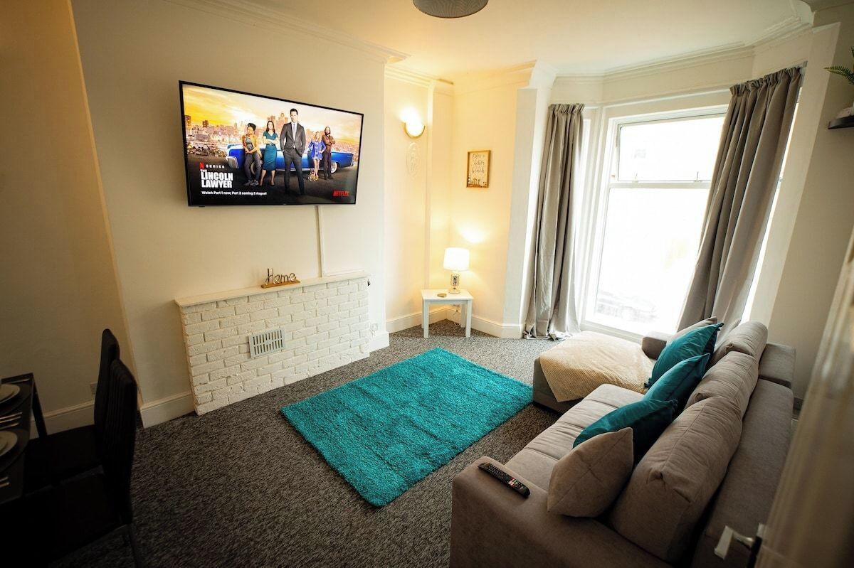 A living room with a flat-screen TV mounted on the wall, a white brick fireplace, a teal rug, a beige sofa with teal cushions, and a window with curtains.