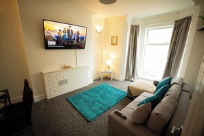 A living room with a flat-screen TV mounted on the wall, a white brick fireplace, a teal rug, a beige sofa with teal cushions, and a window with curtains.