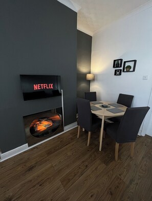 A modern living room with a fireplace, a dining table with chairs, and a flat-screen TV mounted on the wall.