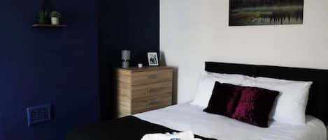 A bedroom with a bed, a bedside table, a nightstand, a wall-mounted shelf with plants, and a painting.