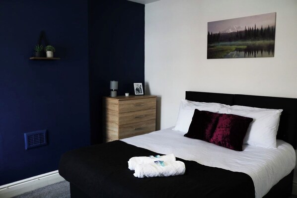 A bedroom with a bed, a bedside table, a nightstand, a wall-mounted shelf with plants, and a painting.